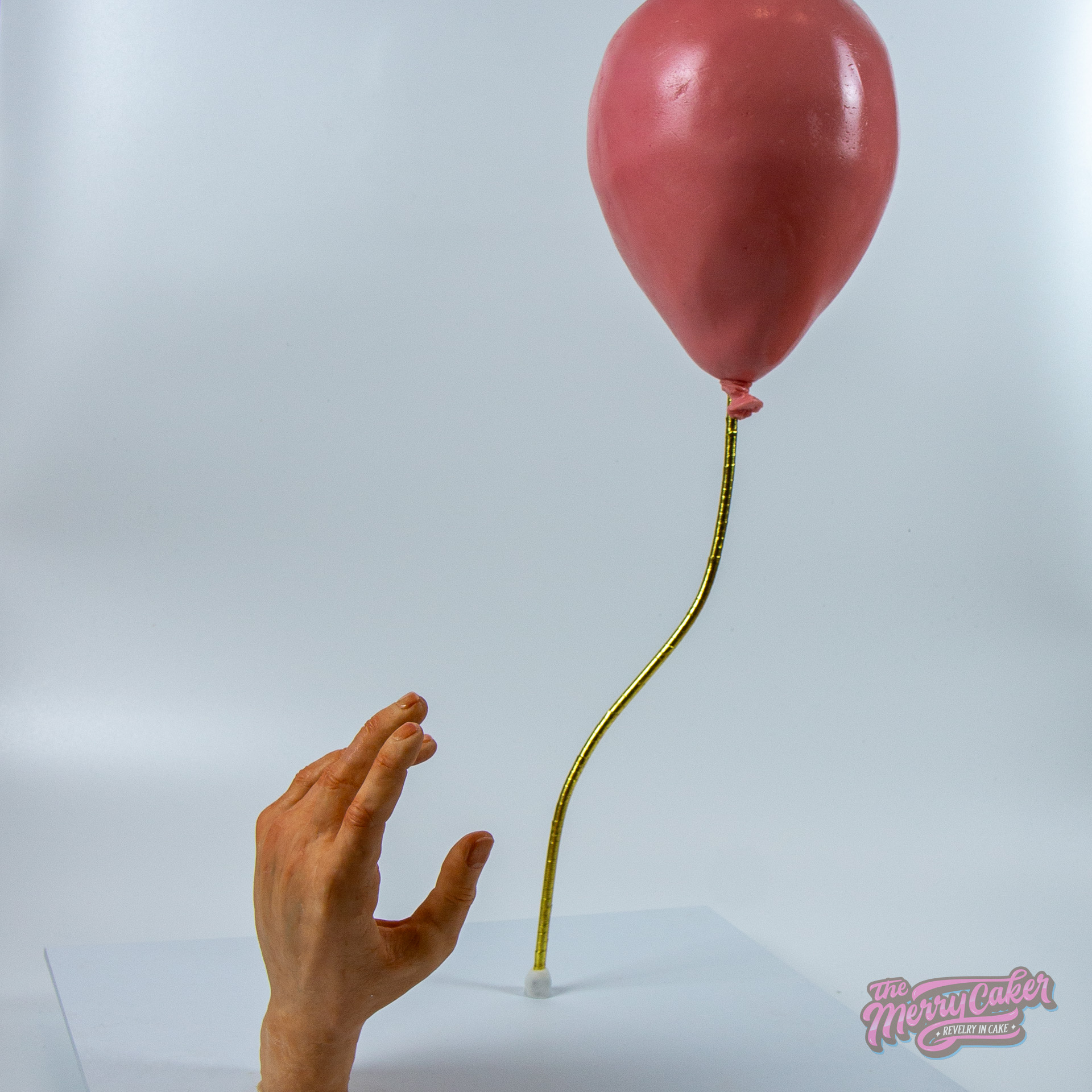 A realistic floating pink balloon cake with a realistic sculpted white chocolate hand letting go of the balloon