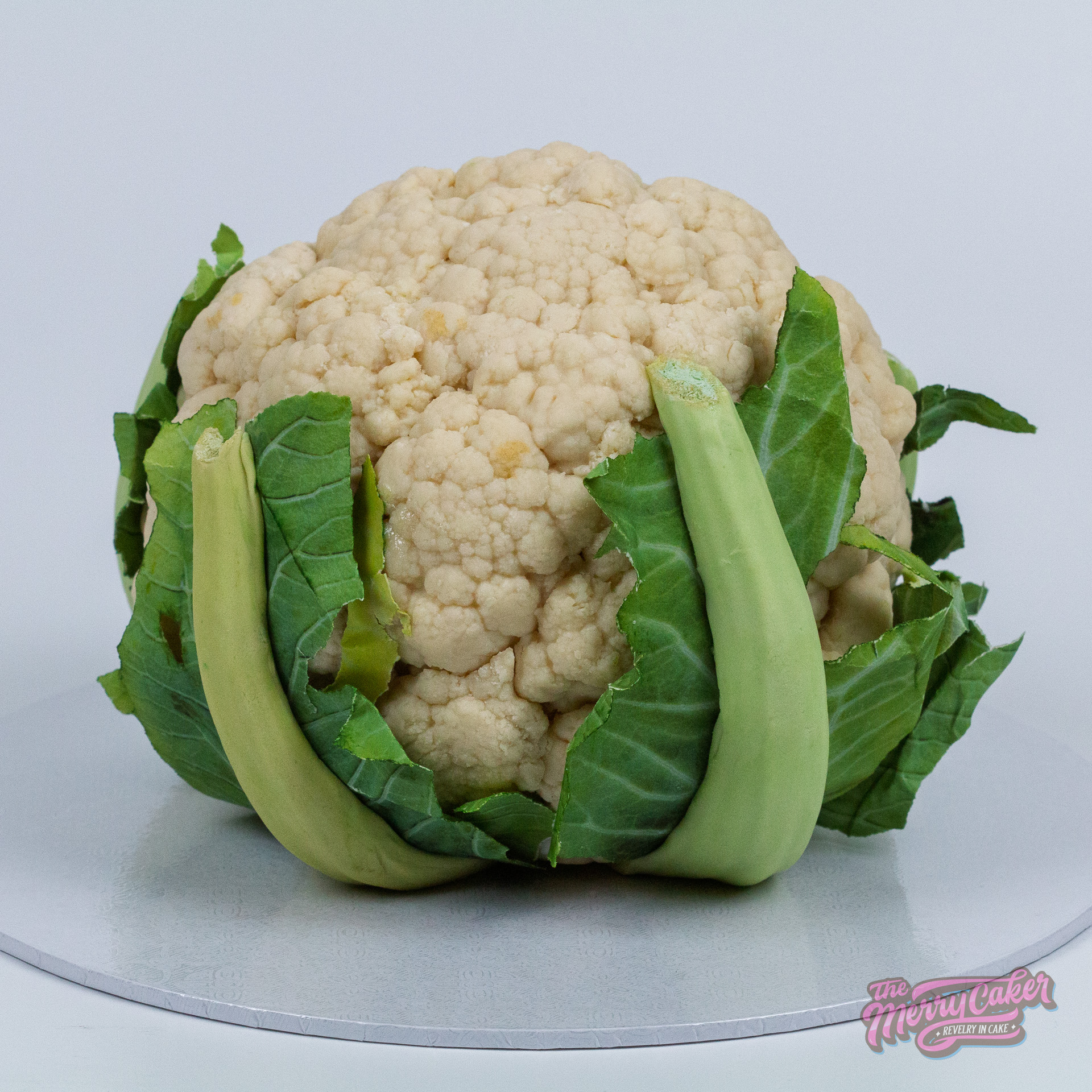 Hyperrealistic cauliflower-shaped cake with detailed green leaves and textured white florets, styled to resemble a real cauliflower, displayed on a silver cake board against a plain light background.
