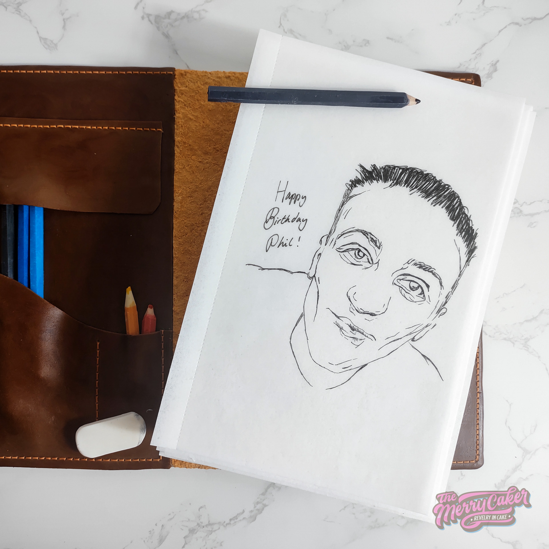 A photograph of a cake that looks like an artist's sketchbook in a leather holder complete with pencils and a rubber.