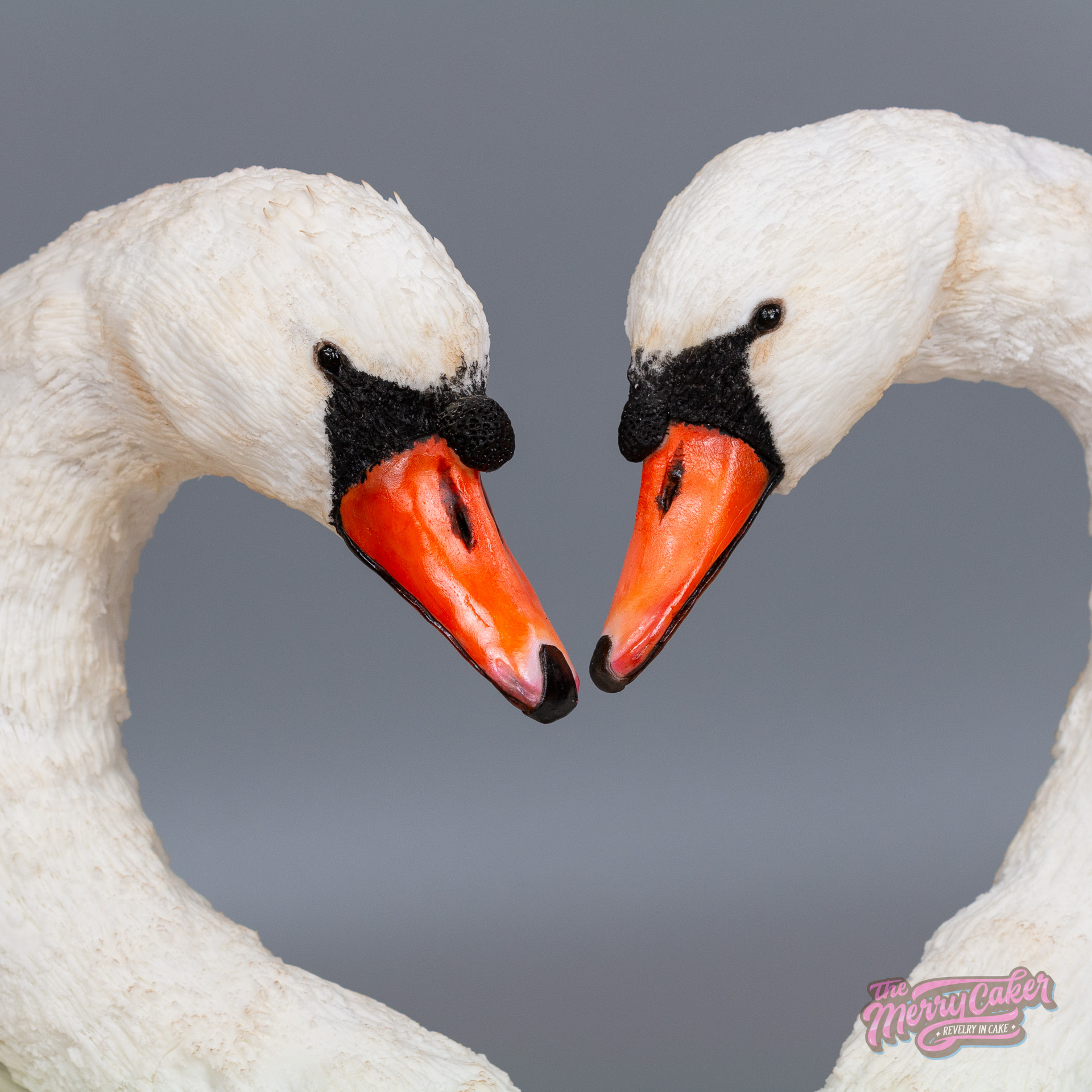 A hyperrealistic swan cake showing two life-size white swans on a cake board. The swans are angled together so their necks make the shape of a love heart. The swan cakes are surrounded by wafer paper flowers and foliage.